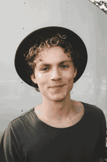 A curly-haired person wearing a black hat stands against a pale wall.