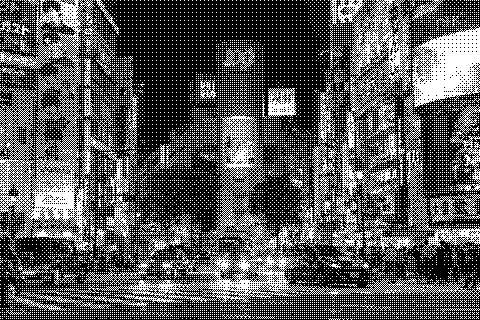 Traffic passes beneath bright signs and neon-lit buildings at a city intersection at night. Dithered grayscale treatment.