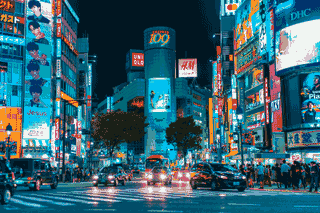 Traffic passes beneath bright signs and neon-lit buildings at a city intersection at night.