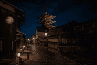 A narrow street leads toward a pagoda at night, with lit buildings along the way.